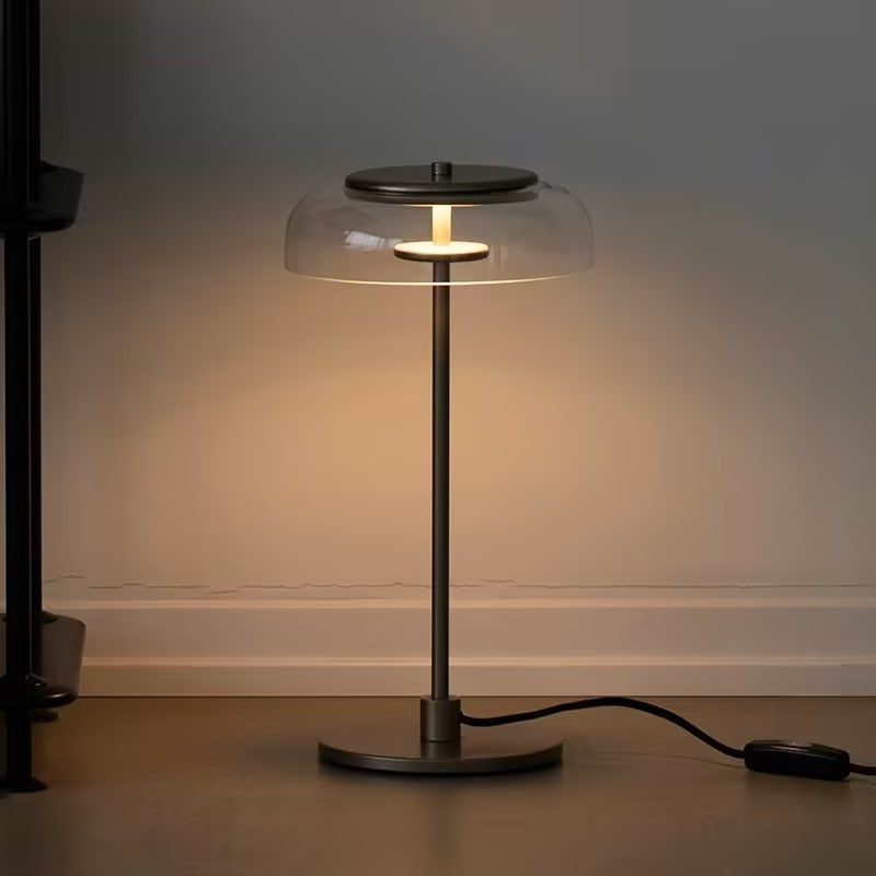 Close view of a Clérachic table lamp with a dark stem and base and a glowing clear dome, standing on a dark floor with its cord switch.