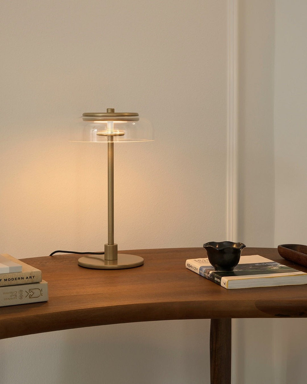 A lit Clérachic table lamp with a brass-toned stem on a curved wooden table, beside a stack of art books and a small black bowl.