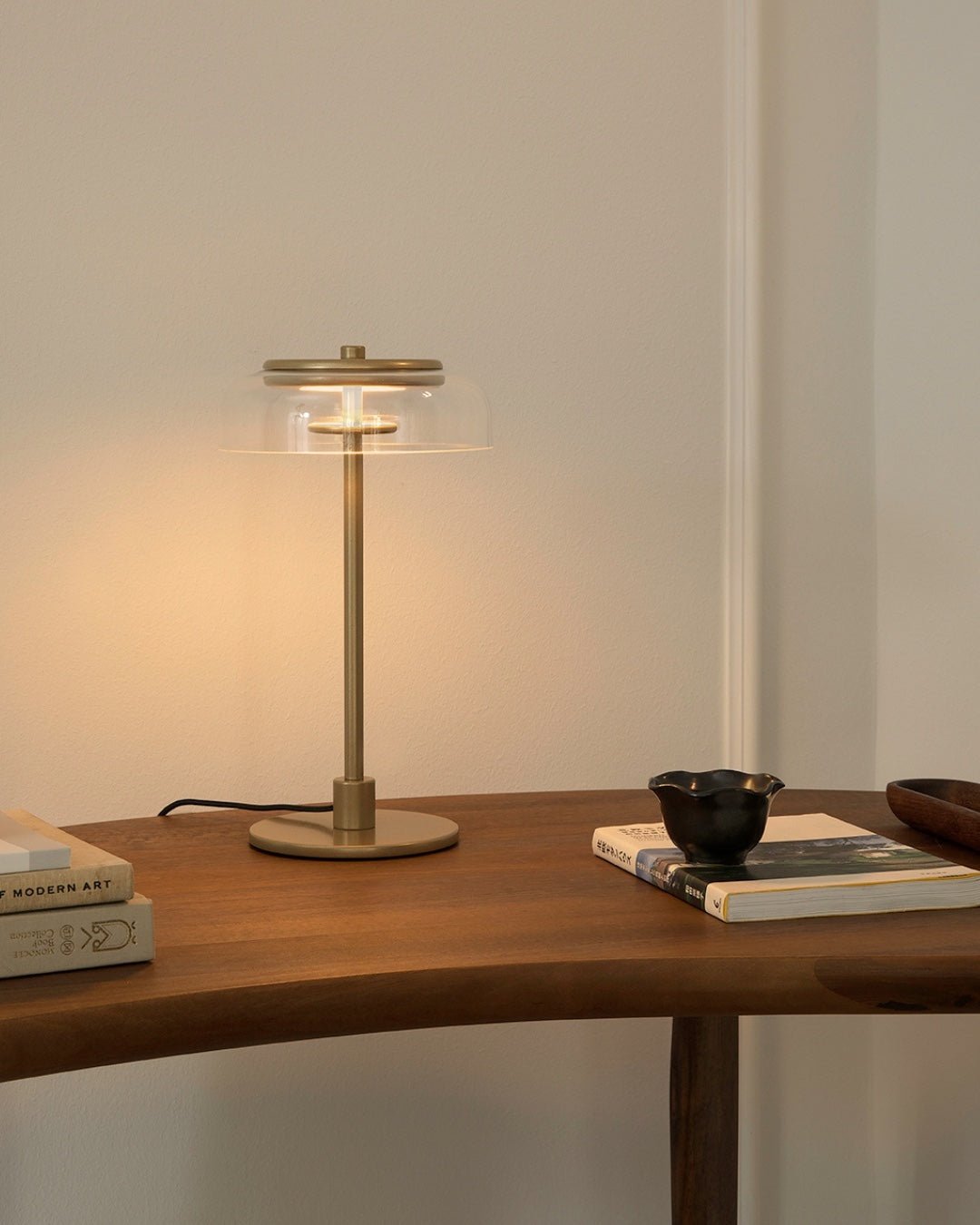 A lit Clérachic table lamp with a brass-toned stem on a curved wooden table, beside a stack of art books and a small black bowl.