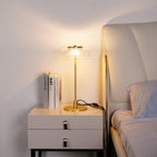 A lit Clérachic table lamp on a white bedside chest next to a bed, with two books standing beside it and a warm glow on the wall.