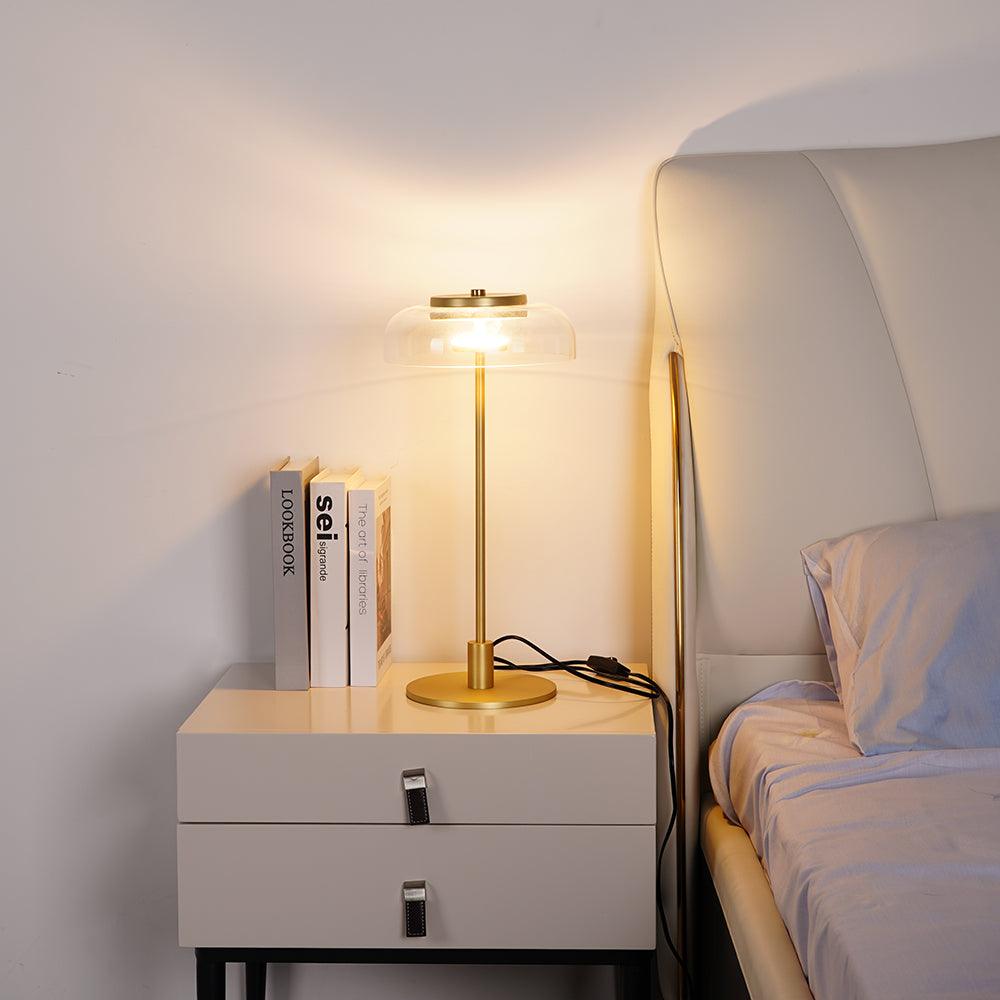 A lit Clérachic table lamp on a white bedside chest next to a bed, with two books standing beside it and a warm glow on the wall.