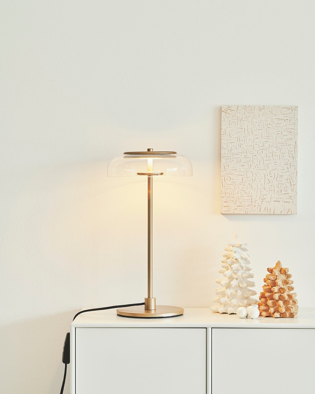 A Clérachic table lamp lit on a white cabinet, with a framed textured artwork and two small ornaments on the wall side.