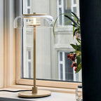 This Clérachic table lamp by a sunny window, with a clear glass dome and a brass-toned stem, next to a dark curtain and a flowering plant.