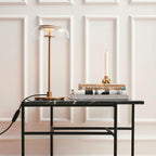 A Clérachic table lamp on a black-framed table with a marble top, in front of white wall panelling, beside a candlestick and books.