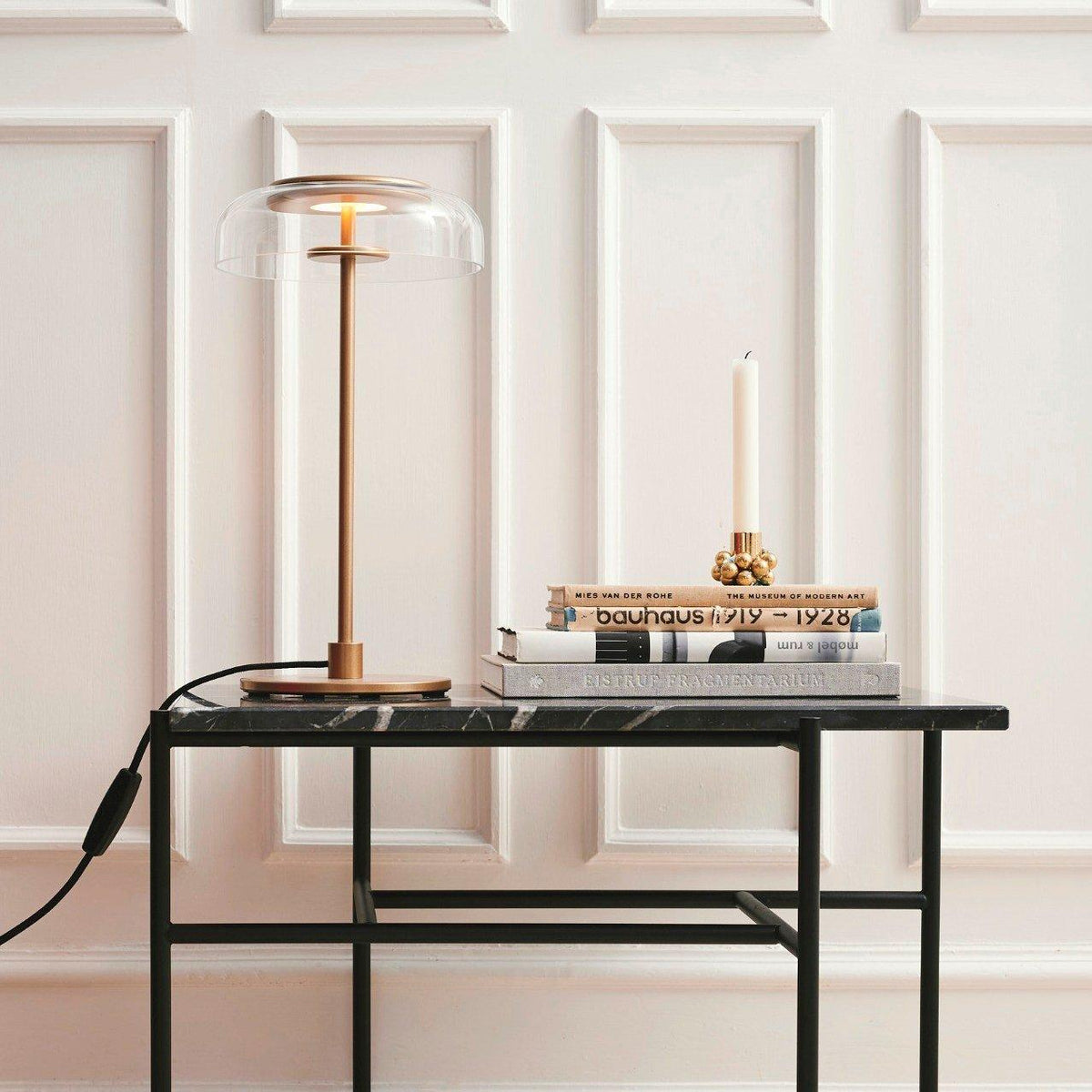 A Clérachic table lamp on a black-framed table with a marble top, in front of white wall panelling, beside a candlestick and books.
