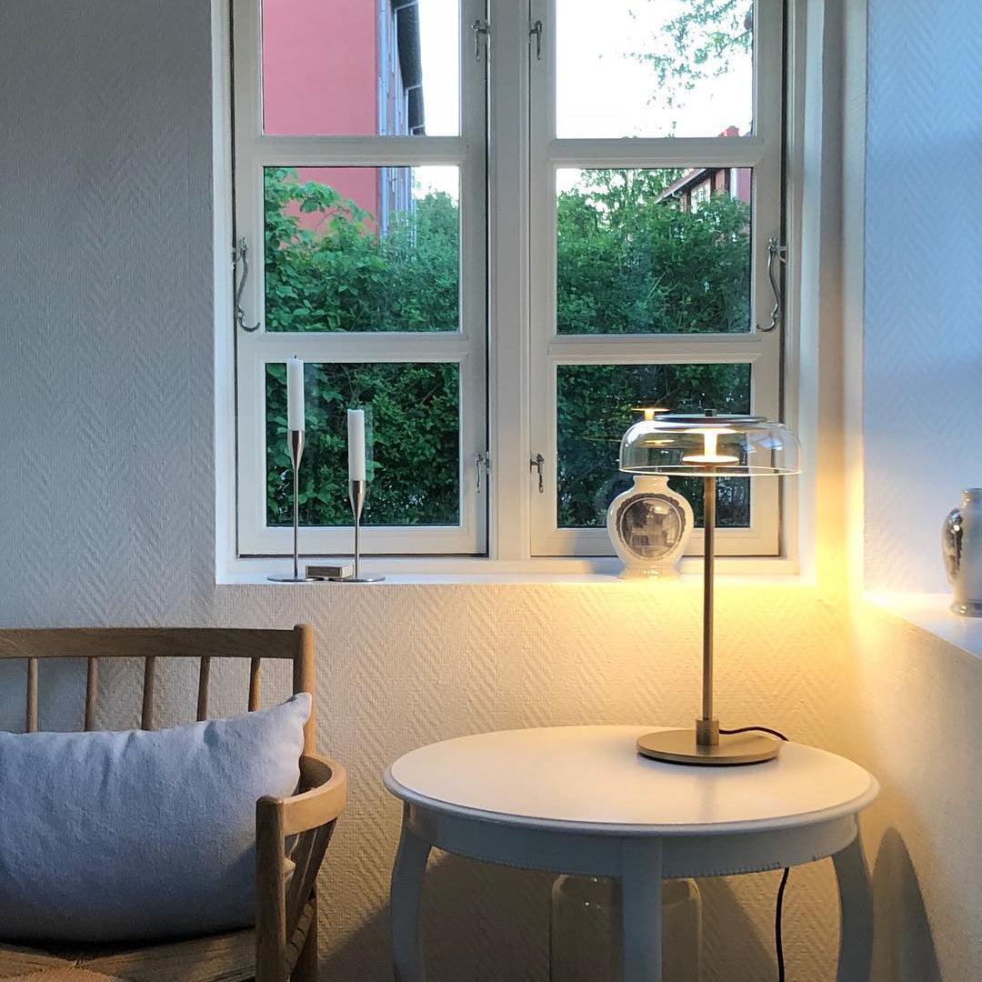 A lit Clérachic table lamp on a small white round table by a window, with candlesticks and a vase on the sill behind it.
