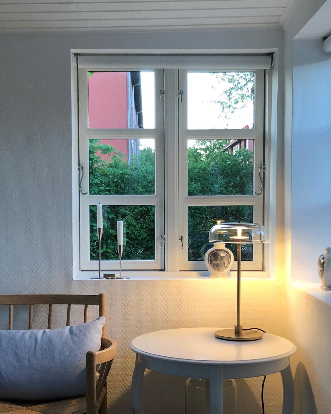A lit Clérachic table lamp on a small round table by a window with two candlesticks, beside a wooden chair and a pale cushion.