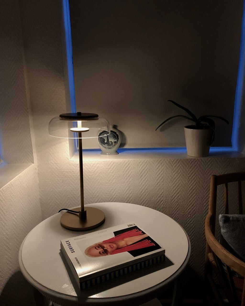 A Clérachic table lamp lit on a round white table with a magazine, in a dim corner with a blue-edged window above.