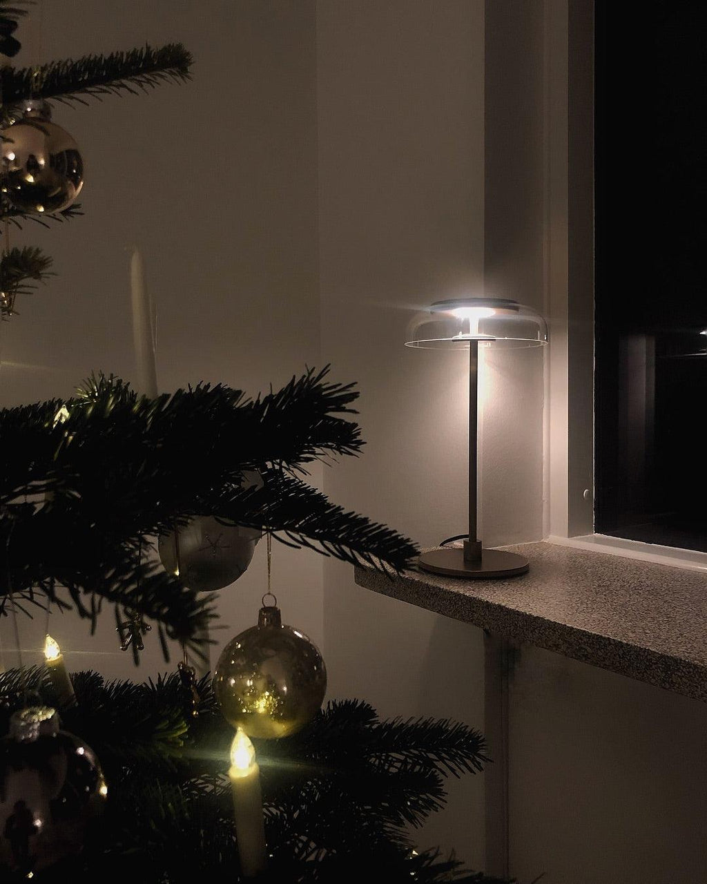A lit Clérachic table lamp on a windowsill beside a decorated fir tree, glowing softly in a dark room.