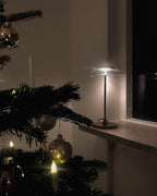 A lit Clérachic table lamp on a windowsill beside a decorated fir tree, glowing softly in a dark room.