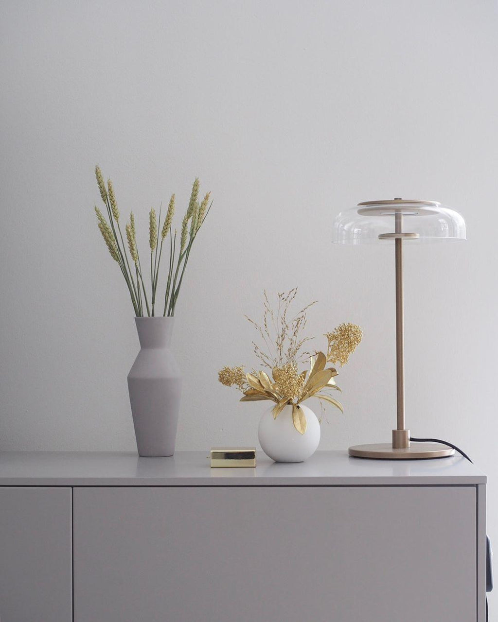 A Clérachic table lamp on a pale sideboard beside a grey vase of dried wheat stems and a small white vase of flowers.