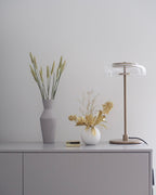 A Clérachic table lamp on a pale sideboard beside a grey vase of dried wheat stems and a small white vase of flowers.