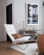 In the corner of a living room with a white lounge chair and a large framed print, a Clérachic table lamp stands on a shelf unit.