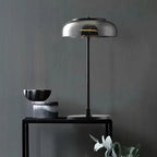 A black-and-smoke-grey Clérachic table lamp on a black console against a dark grey wall, beside a stone bowl and a marble block.