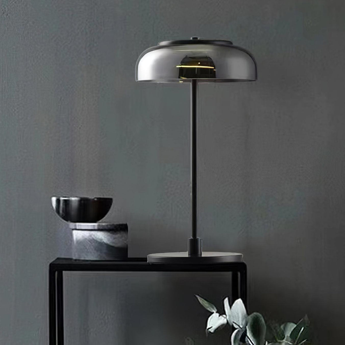 A black-and-smoke-grey Clérachic table lamp on a black console against a dark grey wall, beside a stone bowl and a marble block.