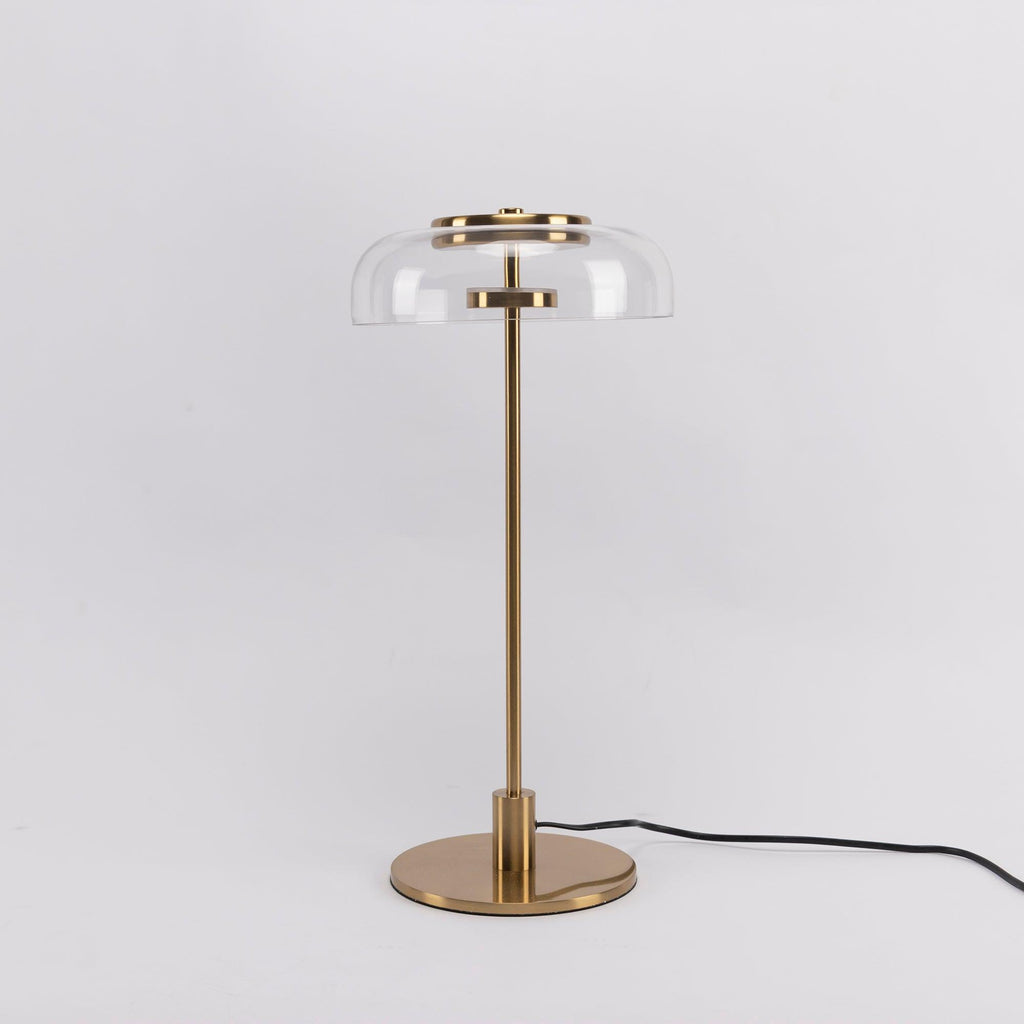 This Clérachic table lamp on a plain pale grey backdrop, with a clear glass dome, a brass stem and a cable running off to the right.