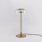 This Clérachic table lamp on a plain pale grey backdrop, with a clear glass dome, a brass stem and a cable running off to the right.