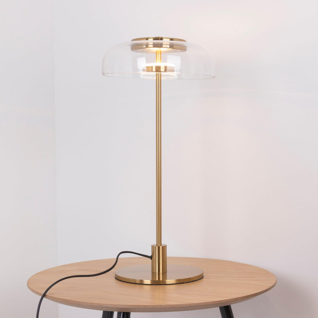 A Clérachic table lamp with a brass stem and a lit glass dome on a round light-oak table, with the cord looping down the side.