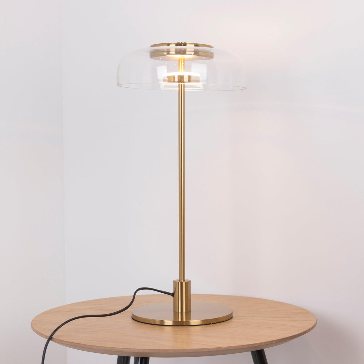 A Clérachic table lamp with a brass stem and a lit glass dome on a round light-oak table, with the cord looping down the side.