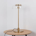 This Clérachic table lamp unlit on a round oak table, with the clear glass dome and brass fittings visible against a pale wall.