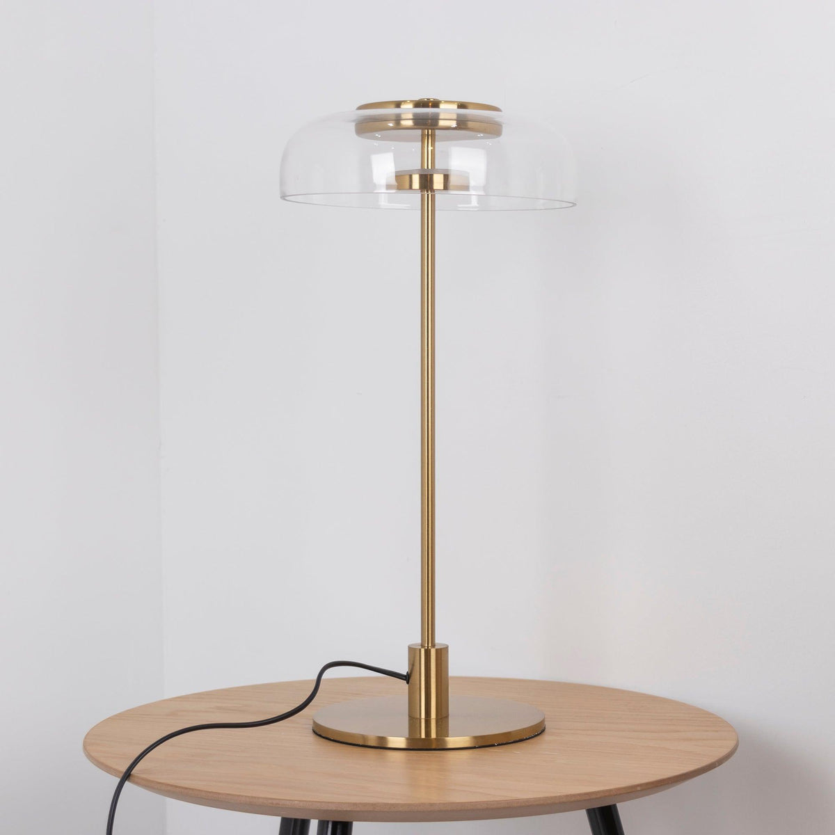 This Clérachic table lamp unlit on a round oak table, with the clear glass dome and brass fittings visible against a pale wall.