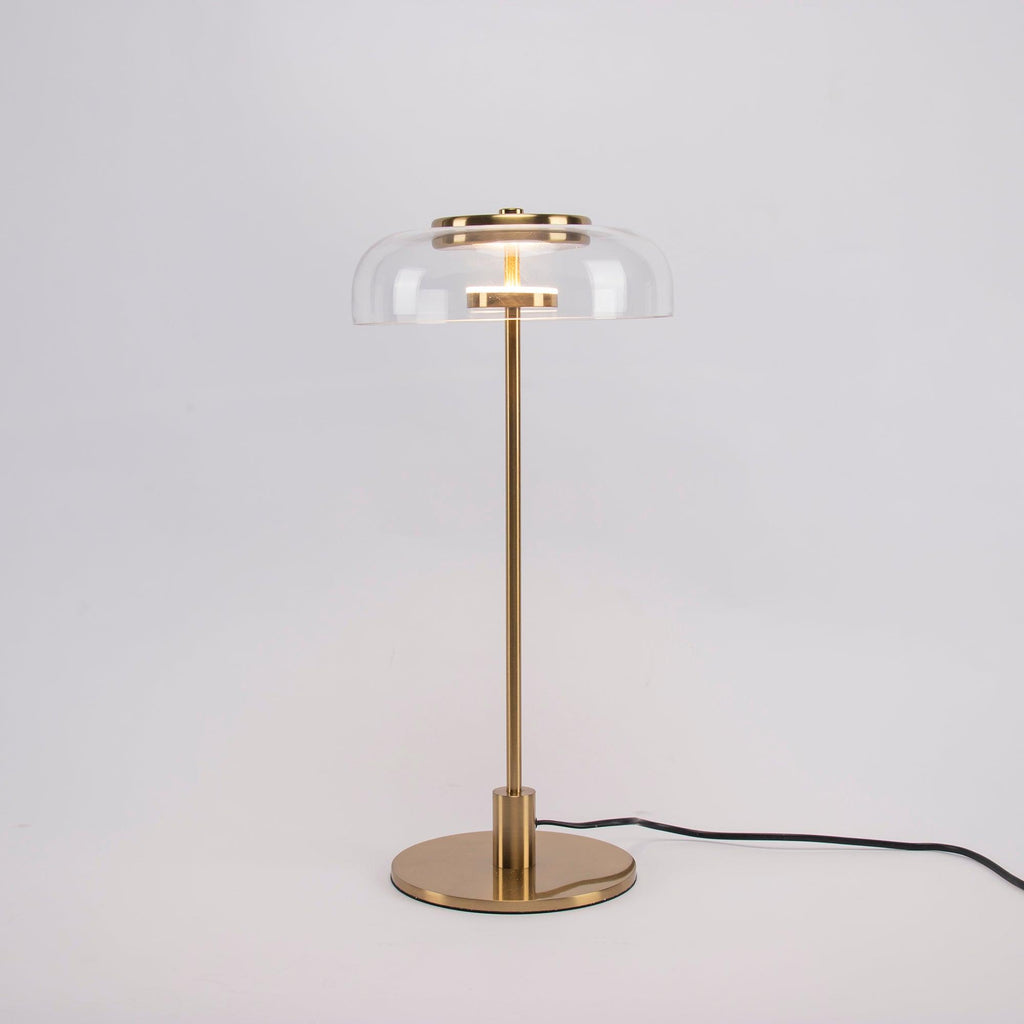 A Clérachic table lamp on a plain grey backdrop, lit, with a short brass stem and a round base and the cord trailing right.