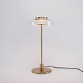 A Clérachic table lamp on a plain grey backdrop, lit, with a short brass stem and a round base and the cord trailing right.
