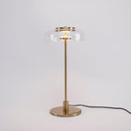 A Clérachic table lamp on a plain grey backdrop, lit, with a short brass stem and a round base and the cord trailing right.