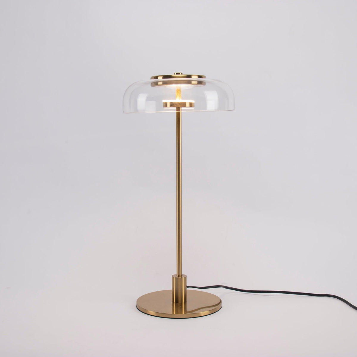 A Clérachic table lamp on a plain grey backdrop, lit, with a short brass stem and a round base and the cord trailing right.