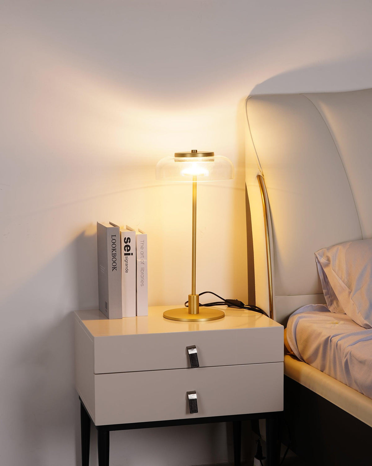 A Clérachic table lamp glowing on a white bedside chest, with a stack of books, a bed and a pale headboard in the background.
