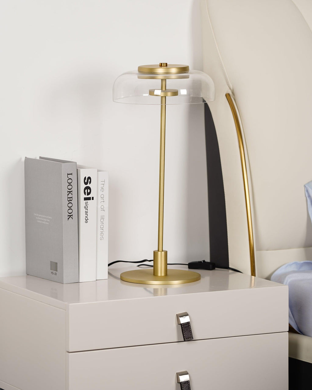 Clérachic table lamp on a bedside chest, unlit, with a clear glass dome and a brass stem, next to three books and a bed.