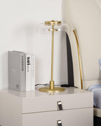 Clérachic table lamp on a bedside chest, unlit, with a clear glass dome and a brass stem, next to three books and a bed.