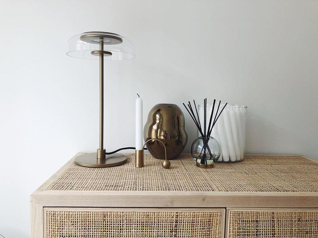 A Clérachic table lamp on a rattan-fronted sideboard, with a taper candle, a brass ornament and a reed diffuser alongside.