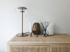 A Clérachic table lamp on a rattan-fronted sideboard, with a taper candle, a brass ornament and a reed diffuser alongside.