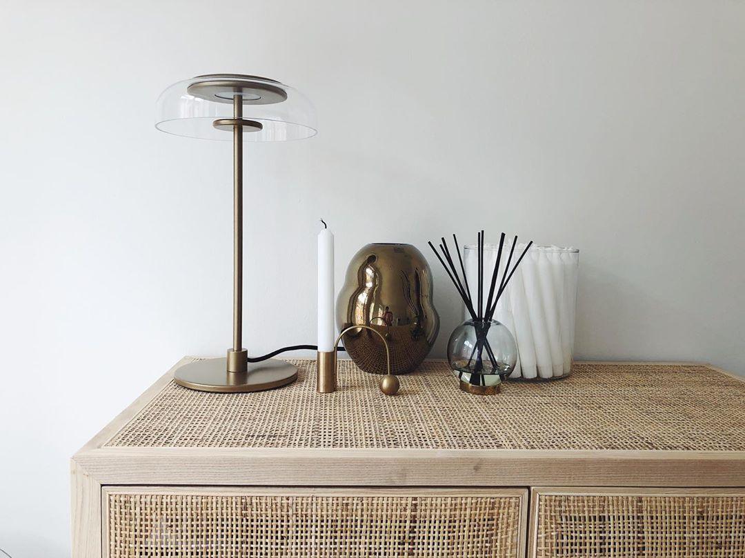 A Clérachic table lamp on a rattan-fronted sideboard, with a taper candle, a brass ornament and a reed diffuser alongside.