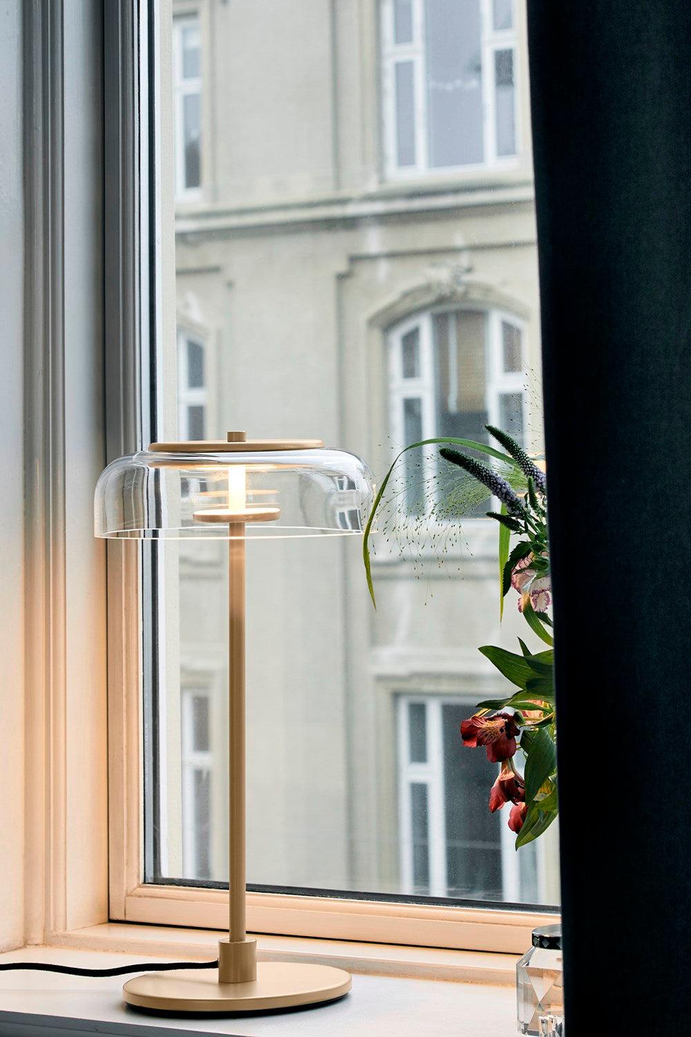 A lit Clérachic table lamp on a windowsill with a dark curtain and a flowering plant, and a pale stone building outside.
