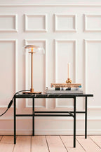 This Clérachic table lamp lit on a marble-topped black table in front of panelled white walls, beside a taper candle and some books.