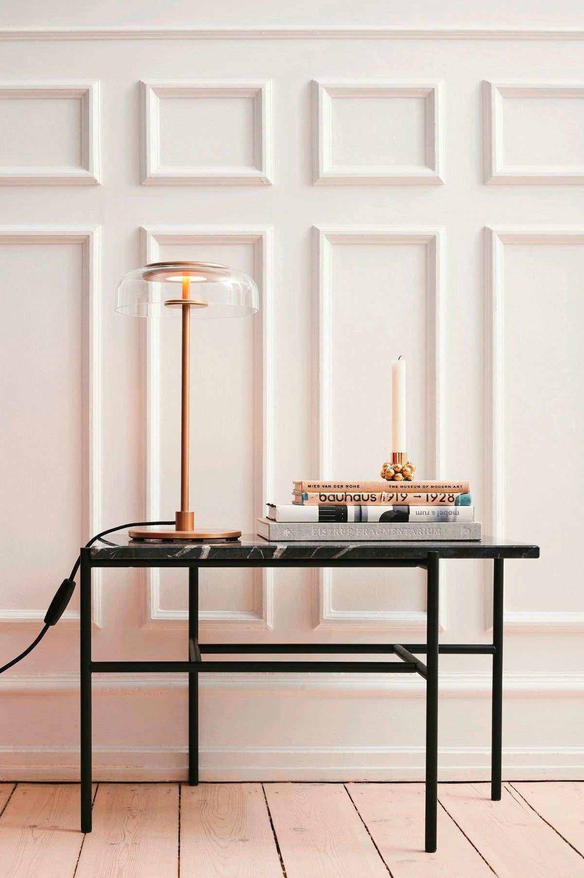 This Clérachic table lamp lit on a marble-topped black table in front of panelled white walls, beside a taper candle and some books.
