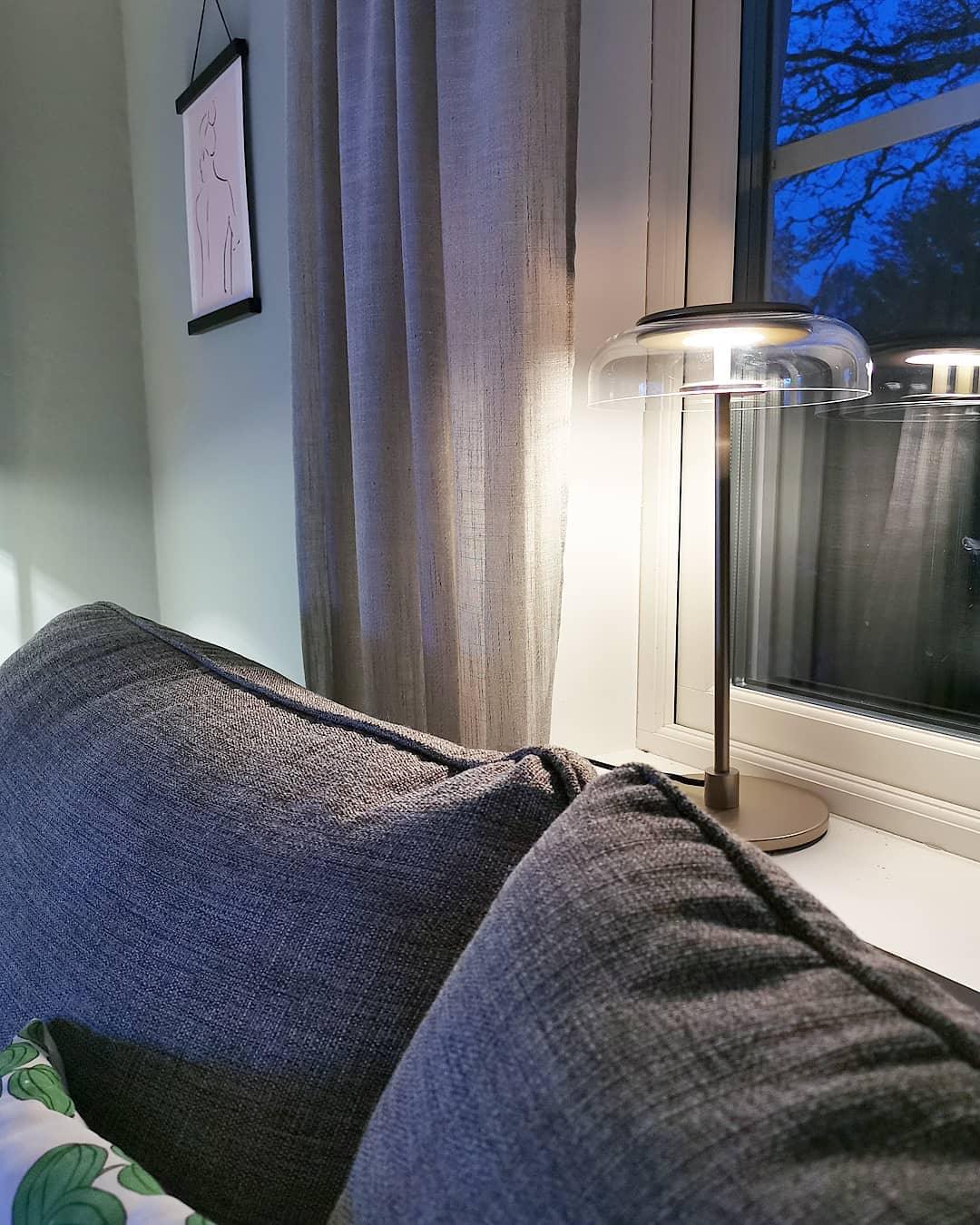 In a blue-lit evening room, a Clérachic table lamp glows on a windowsill behind grey cushions, with a curtain and framed sketch above.