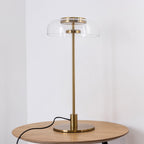 A Clérachic table lamp on a round oak table against a white wall, with a clear dome, a brass cap and a brass stem.