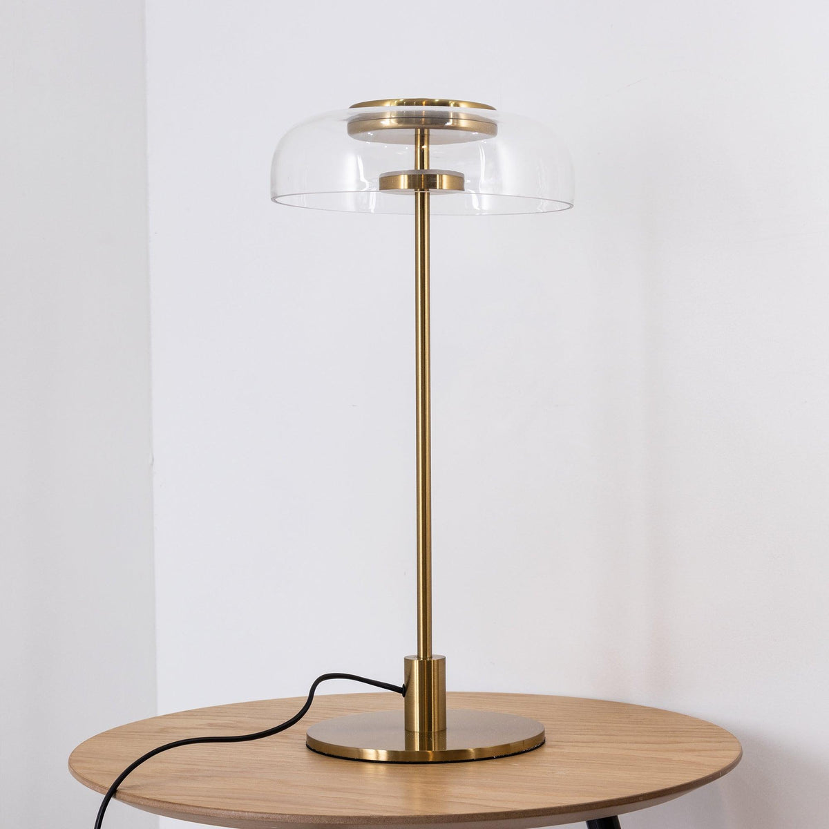 A Clérachic table lamp on a round oak table against a white wall, with a clear dome, a brass cap and a brass stem.