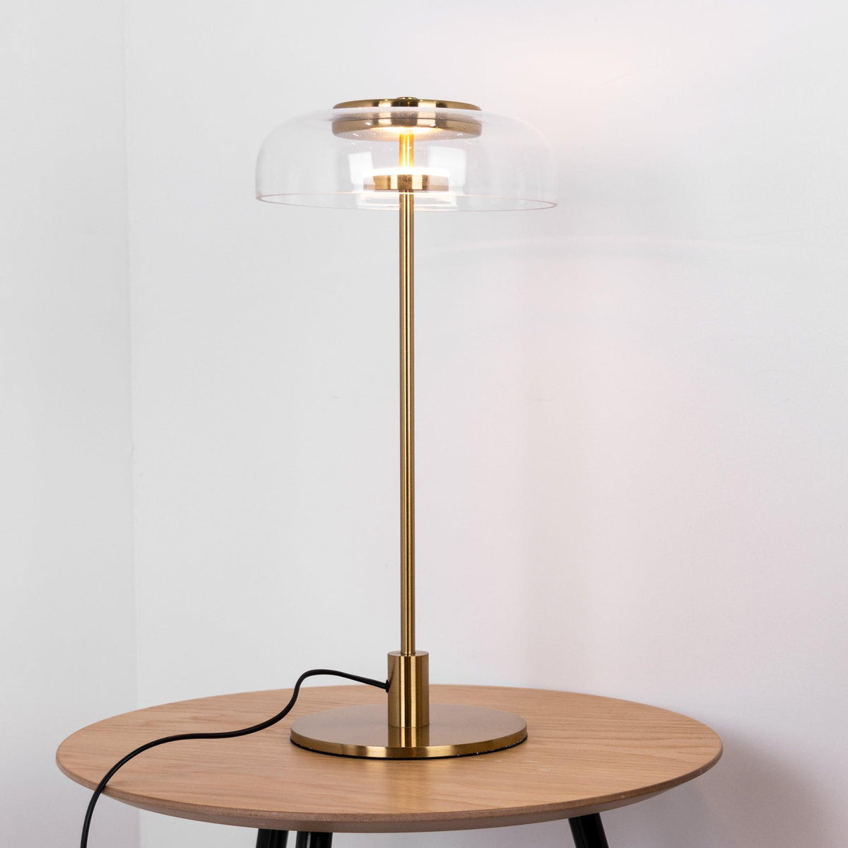 A brass-toned Clérachic table lamp lit on a round oak table, with its cord looping behind it and a pale wall behind.