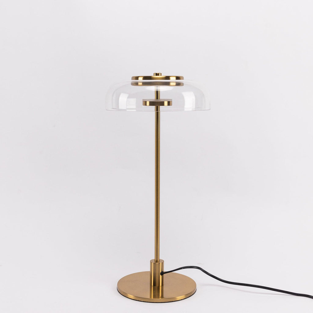 A Clérachic table lamp with a clear glass dome and brass stem on a pale grey backdrop, shown full length with its cord.