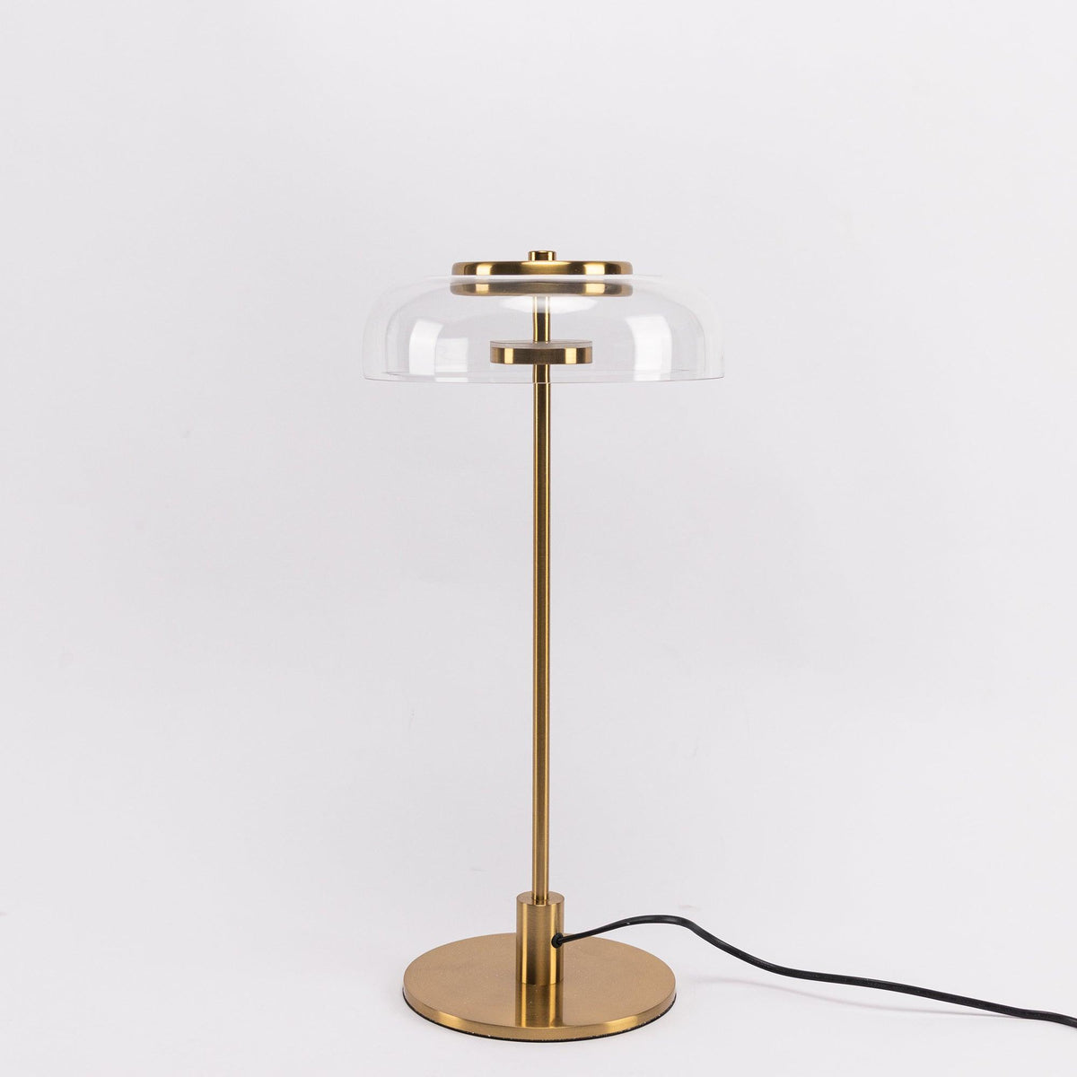 A Clérachic table lamp with a clear glass dome and brass stem on a pale grey backdrop, shown full length with its cord.
