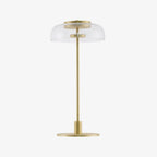 A Clérachic table lamp with a clear dome and a slim stem on a white background, with a flat, round brass-toned base.