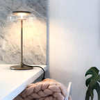 A lit Clérachic table lamp on a white shelf beside a marble wall, with a chunky pink knitted blanket below.