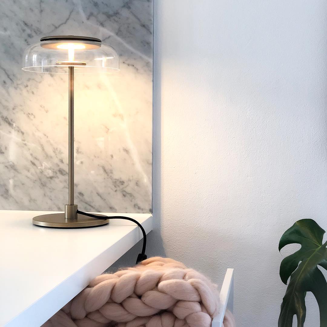 A lit Clérachic table lamp on a white shelf beside a marble wall, with a chunky pink knitted blanket below.