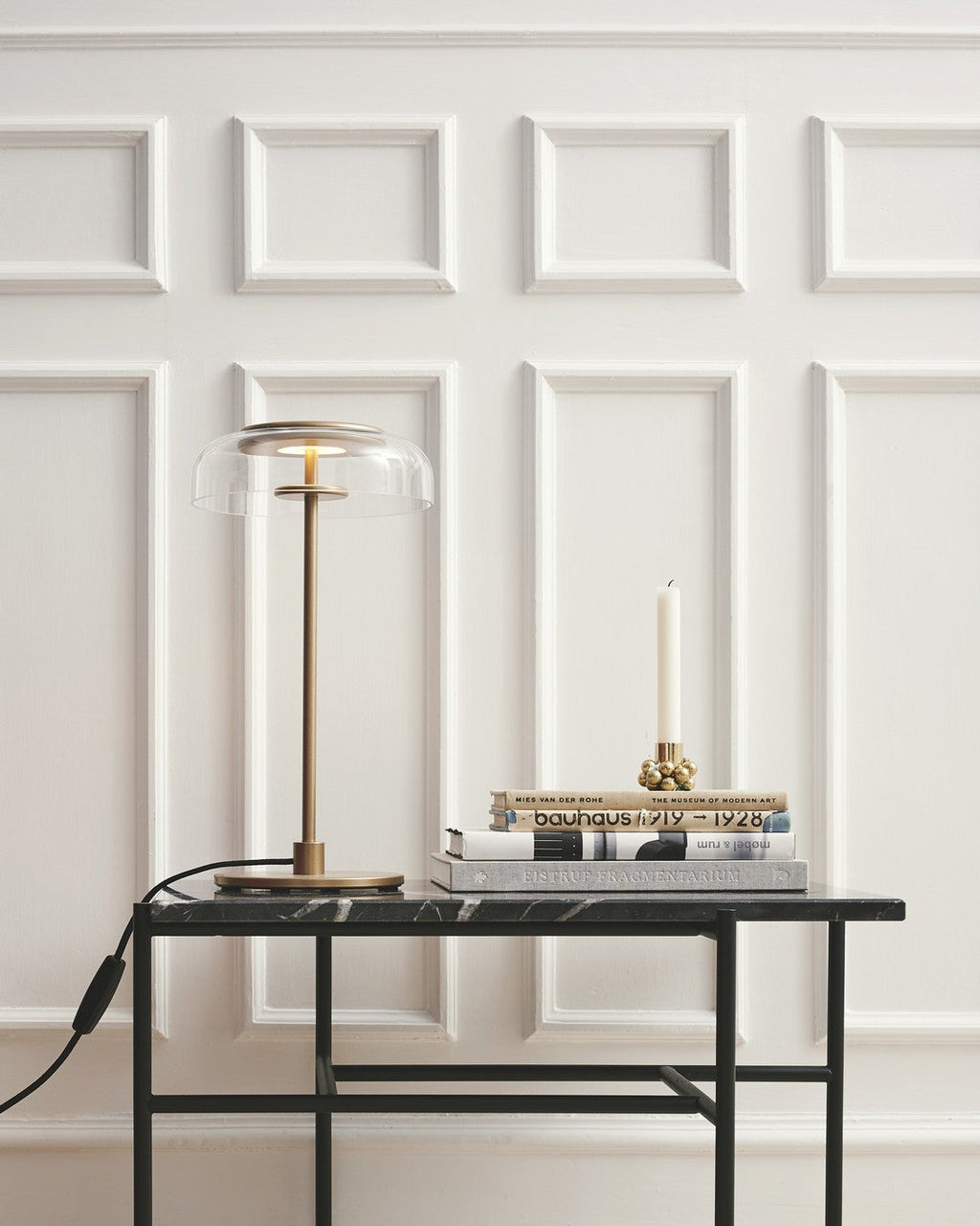 A Clérachic table lamp on a black-framed marble-topped table in front of white wall panelling, unlit, beside a candle and books.
