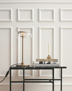 A Clérachic table lamp on a black-framed marble-topped table in front of white wall panelling, unlit, beside a candle and books.