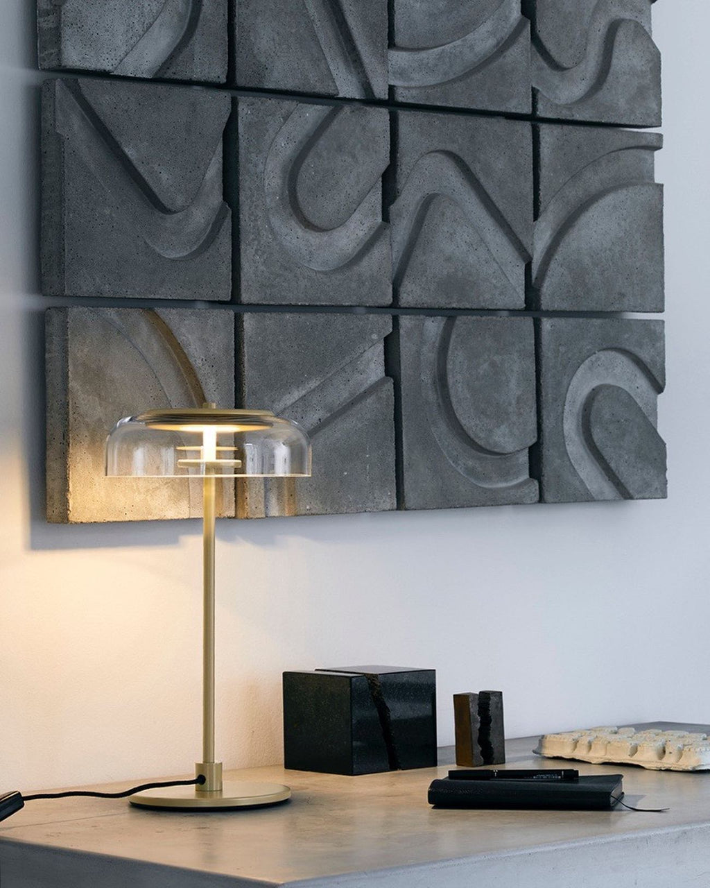 A lit Clérachic table lamp with a glass dome on a grey surface in front of a carved stone wall panel, with dark ornaments beside it.